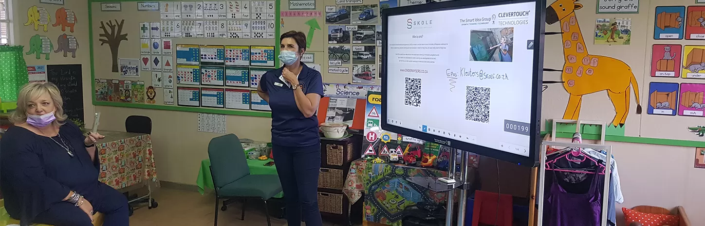 Teacher in a face mask standing next to a IMPACT Plus display in a primary school classroom